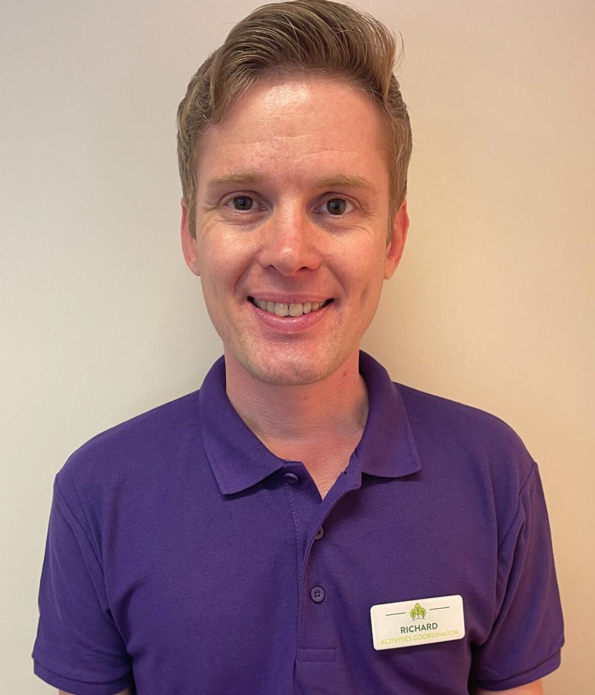man with blonde hair smiling in a purple polo shirt